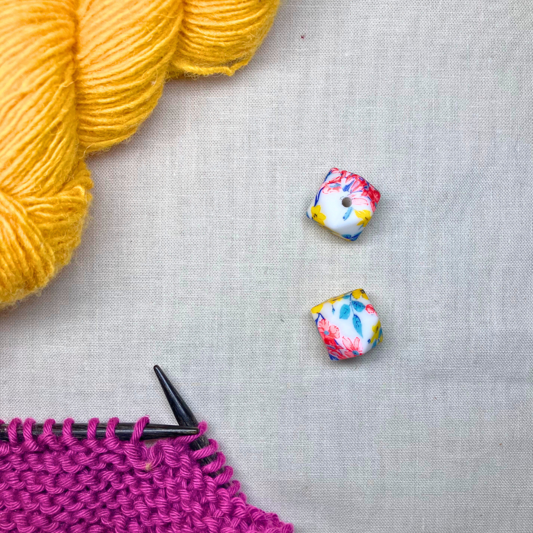 Colourful, geometric stitch stoppers on a lightly textured surface framed by yellow & purple yarn