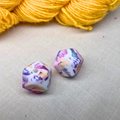 Two colourful, geometric stitch stoppers on a lightly textured surface with yellow yarn in the background