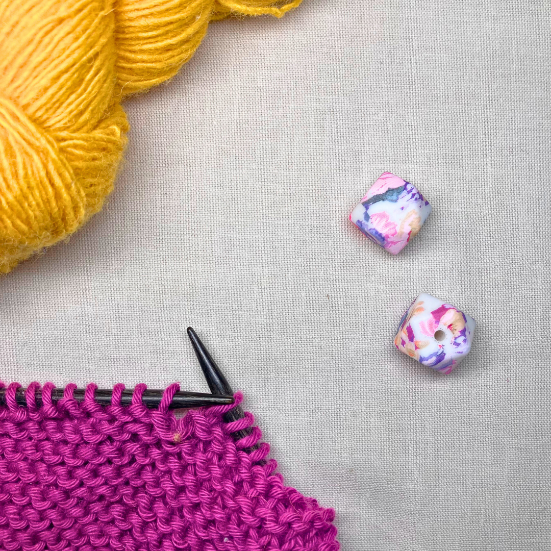 Colourful, geometric stitch stoppers on a lightly textured surface, framed yellow & purple yarn