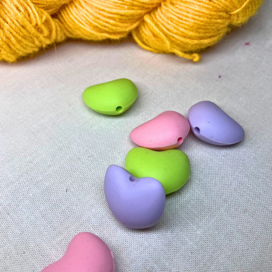 Pastel-coloured, heart-shaped stitch stoppers on a lightly textured surface with yellow yarn in the background