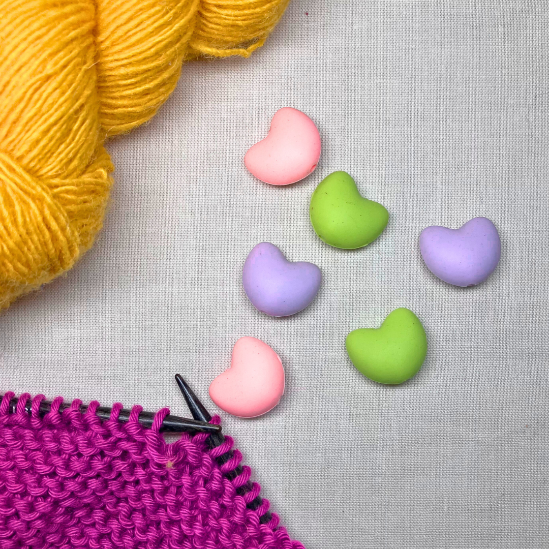 Pastel-coloured, heart-shaped stitch stoppers on a lightly textured surface framed by yellow & purple yarn