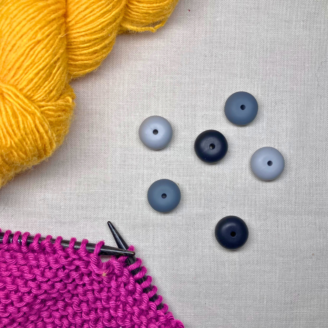 Monochrome, geometric stitch stoppers on a lightly textured surface, framed by yellow & purple yarn