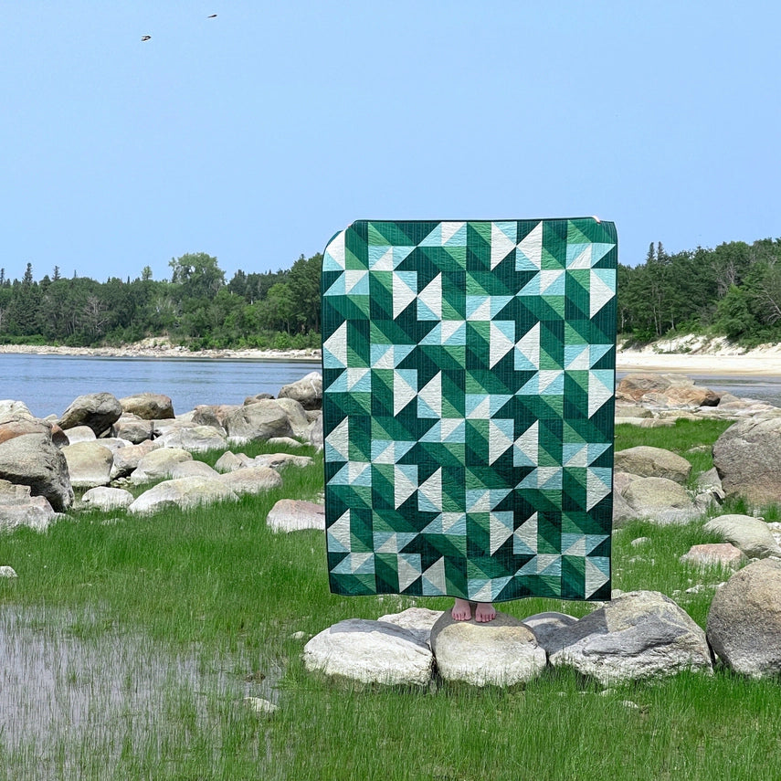 Green and white geometric quilt displayed outdoors by a body of water with rocks and grass.