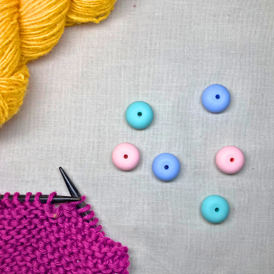 Pastel-coloured, oval stitch stoppers on a lightly textured surface, framed by yellow & purple yarn