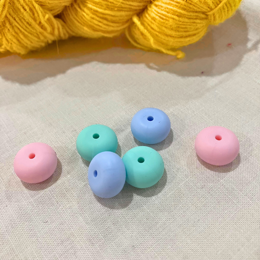 Pastel-coloured, oval stitch stoppers on a lightly textured surface with yellow yarn in the background