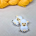 Two white sheep-shaped stitch stoppers with black accents on a lightly textured background with yellow yarn in the background