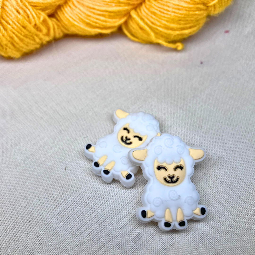 Two white sheep-shaped stitch stoppers with black accents on a lightly textured background with yellow yarn in the background