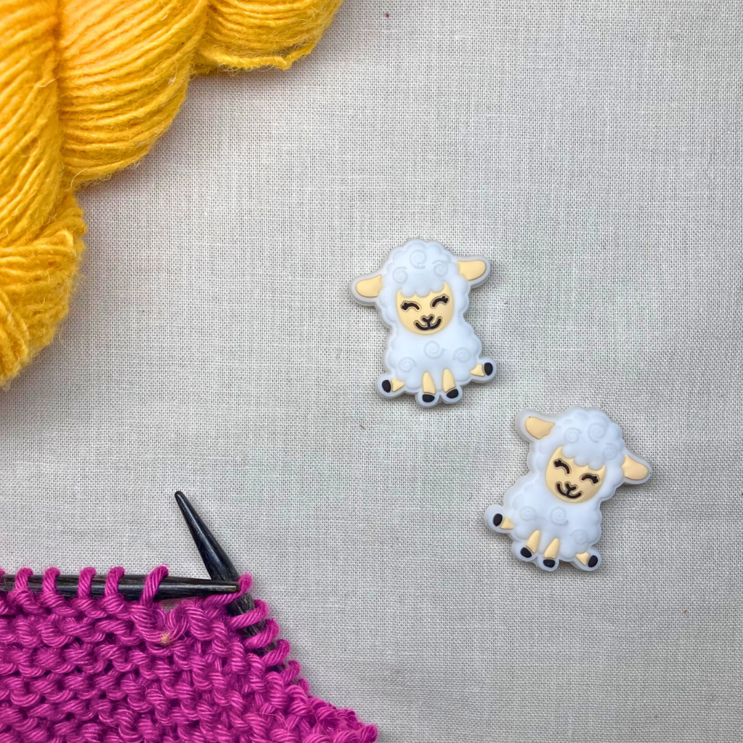 Two white sheep-shaped stitch stoppers with black accents on a lightly textured background, framed by yellow & purple yarn