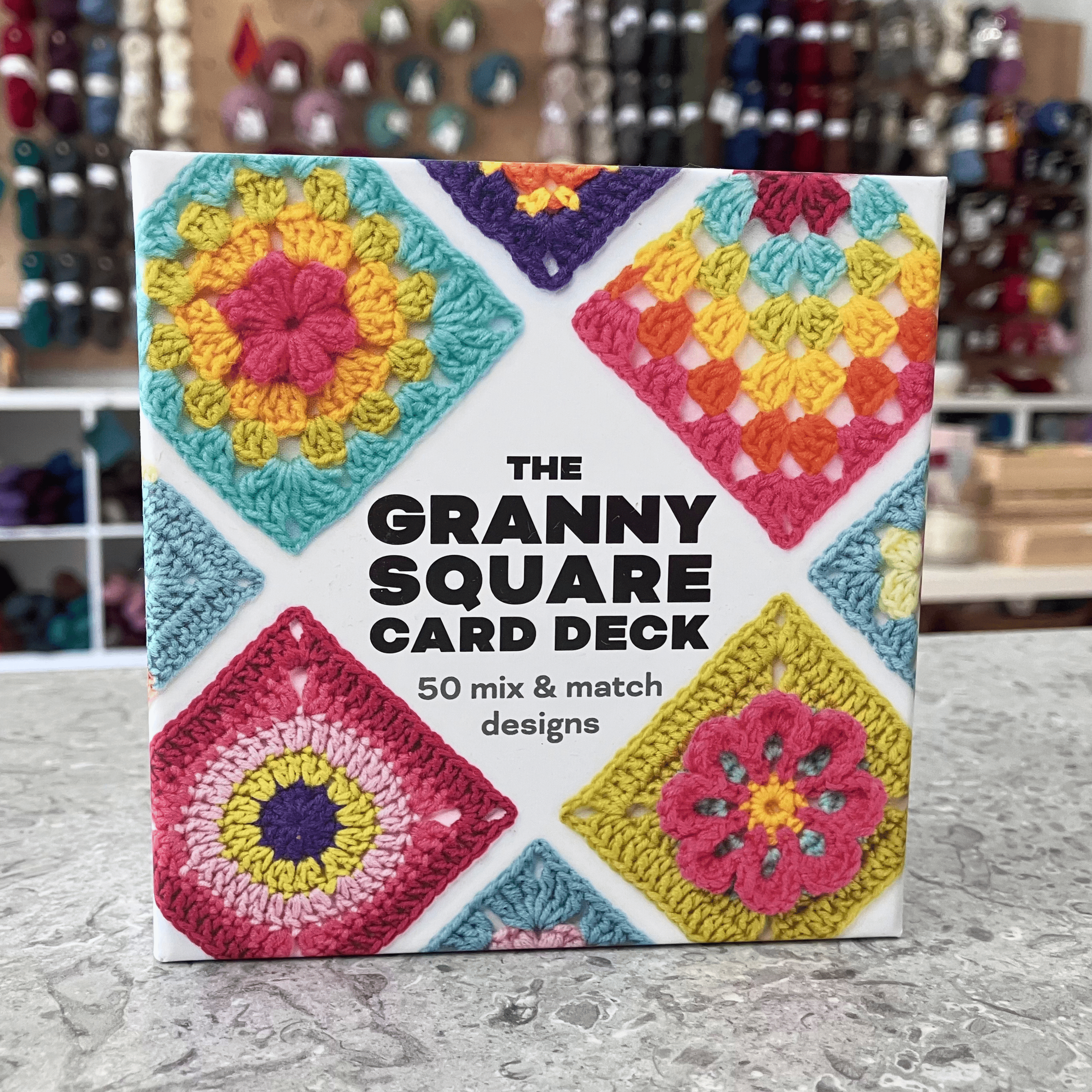 Granny Square Card Deck — Search Press 50-card crochet granny square reference with booklet. Craft shop background (blurred).