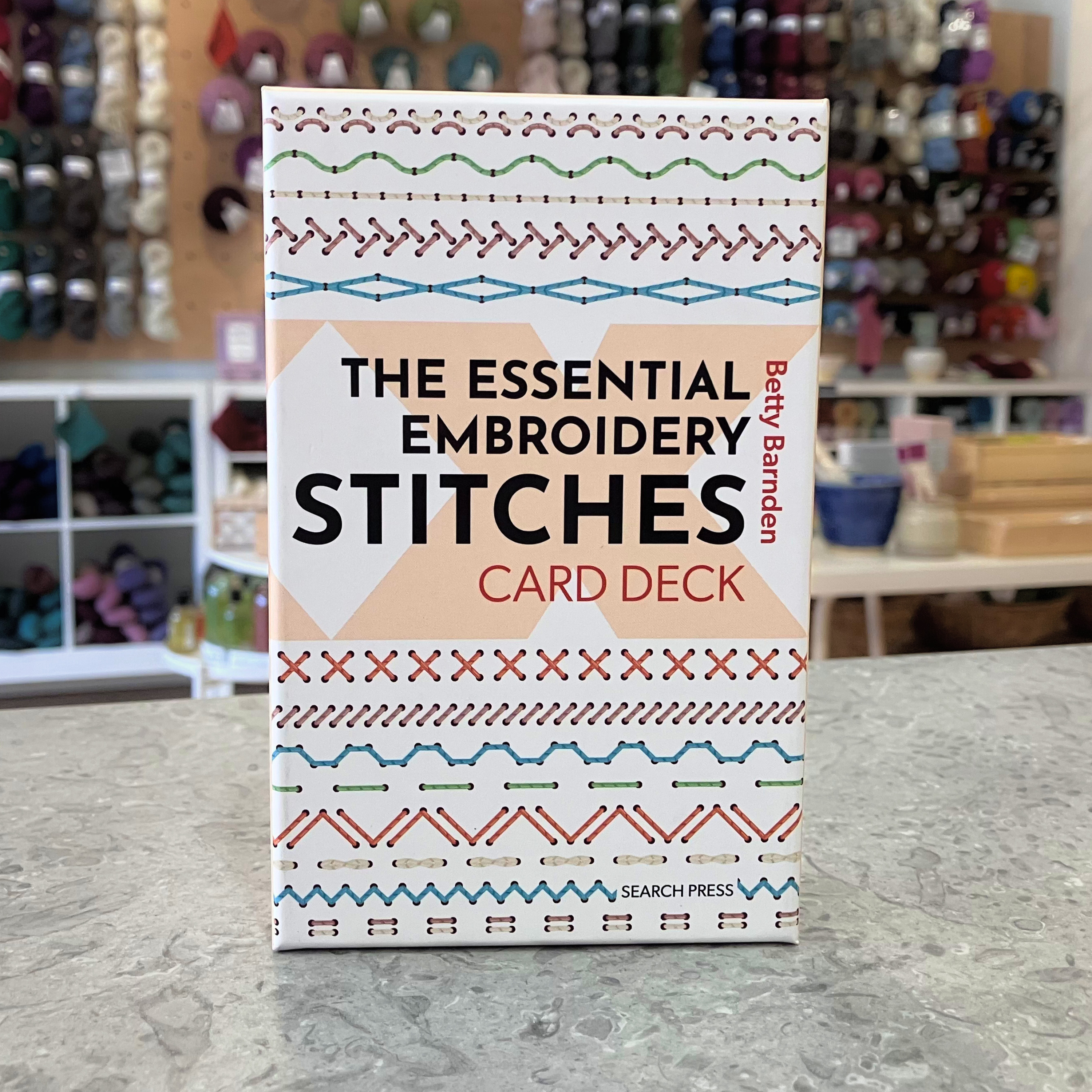 Essential Embroidery Stitches Card Deck by Betty Barnden — 50-card embroidery stitch reference with booklet. Craft shop background (blurred).