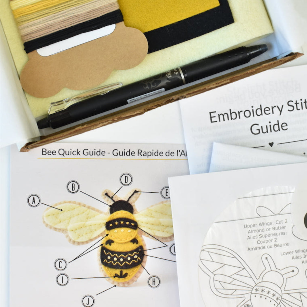 Embroidery kit contents arranged in a box with a bee-themed quick guide, embroidery stitch guide, thread spool, yellow felt sheet, black pen, and other small accessories on a white background.