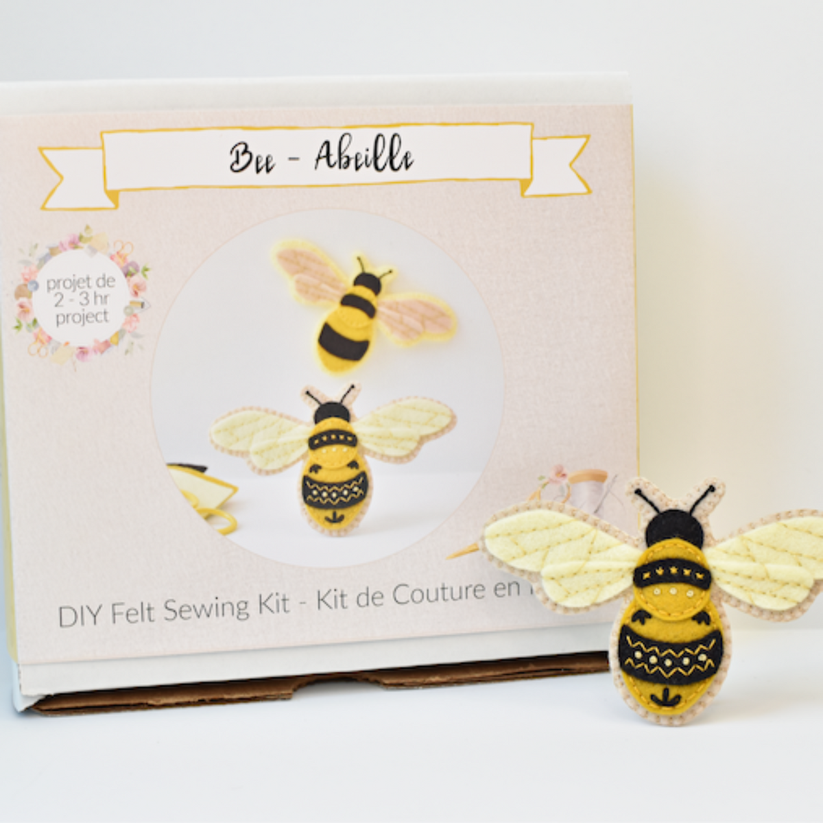 A DIY felt sewing kit box labeled 'Bee - Abeille' stands next to a small felt bee craft with yellow and black patterned body and pale yellow wings on a white background.