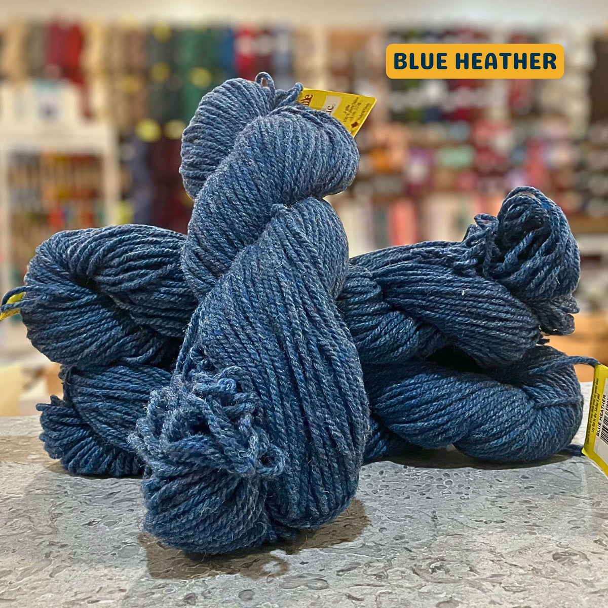 113g dark denim (blue heather) Canadian wool bulky skein, Briggs & Little yarn, craft shop background (blurred)