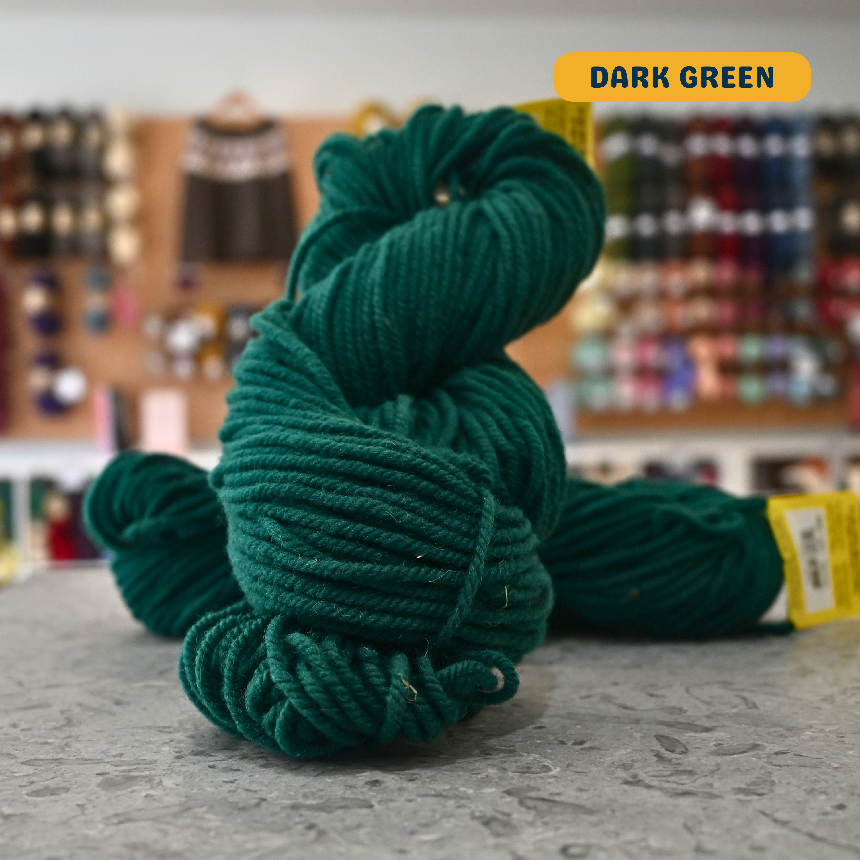 113g dark green Canadian wool bulky skein, Briggs & Little yarn, craft shop background (blurred)
