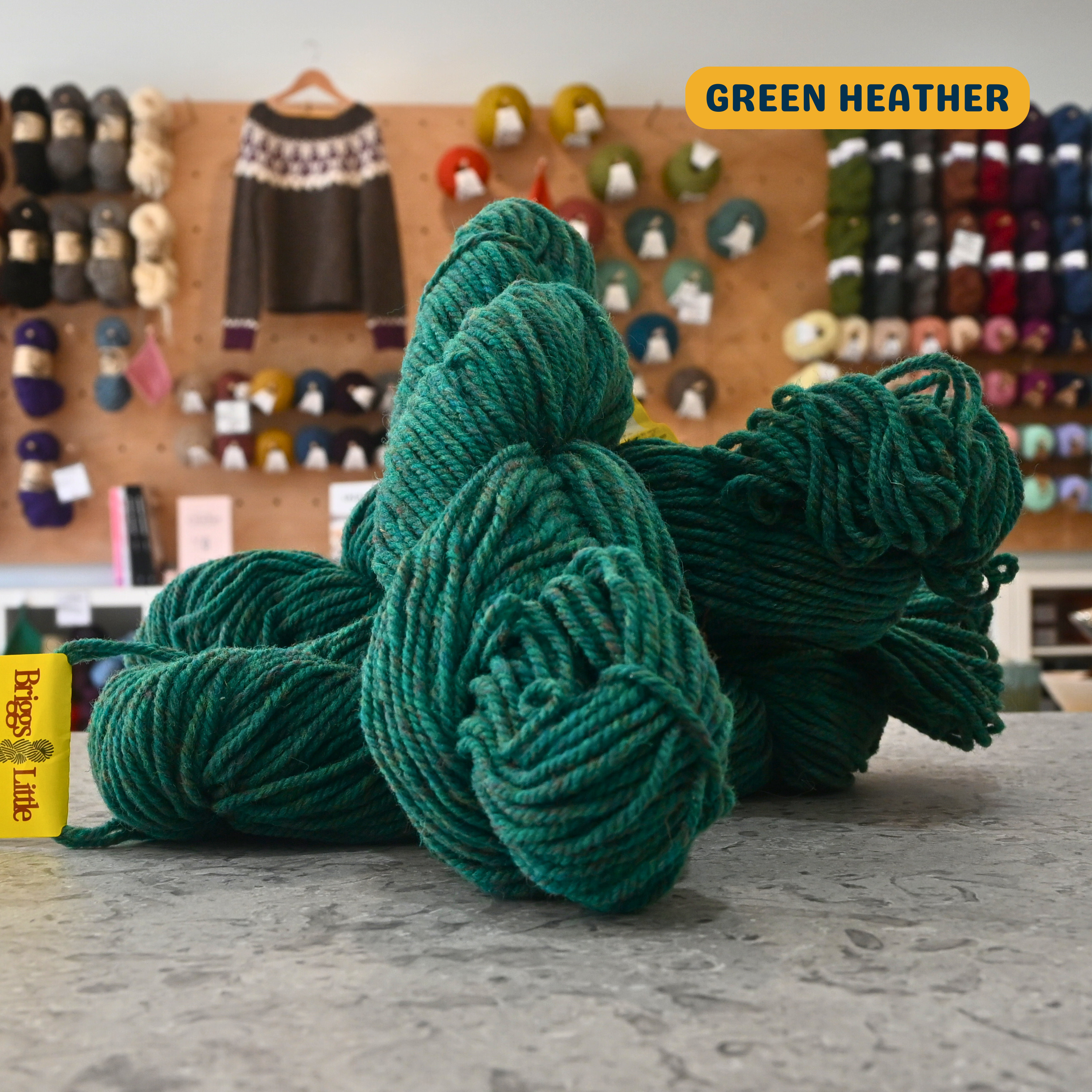 113g heathered green Canadian wool bulky skein, Briggs & Little yarn, craft shop background (blurred)

