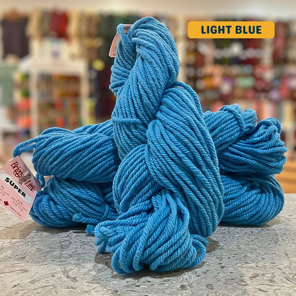113g bright blue (light blue) Canadian wool super bulky skein, Briggs & Little yarn, craft shop background (blurred)