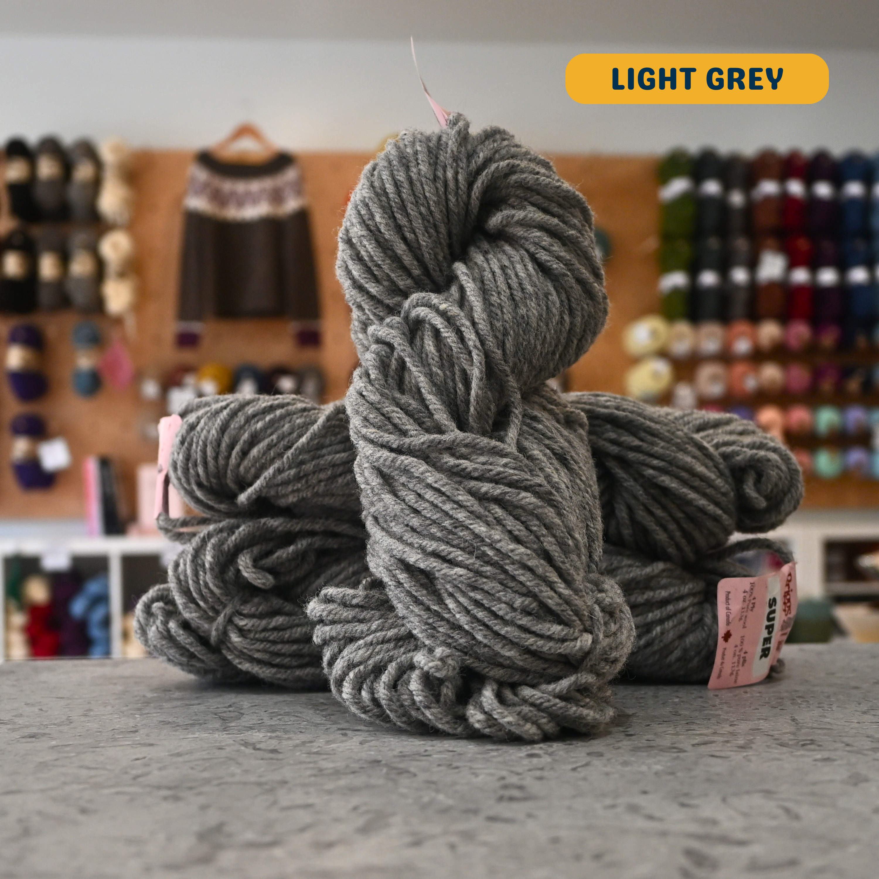 113g light grey Canadian wool super bulky skein, Briggs & Little yarn, craft shop background (blurred)
