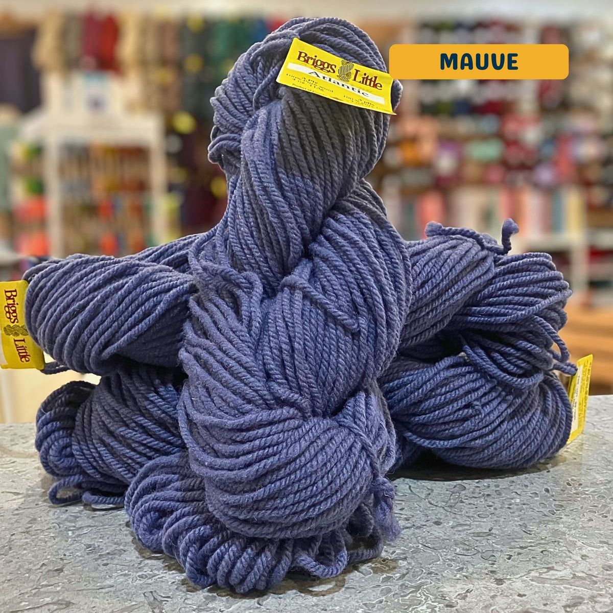 113g dusty blue-purple (mauve) Canadian wool bulky skein, Briggs & Little yarn, craft shop background (blurred)