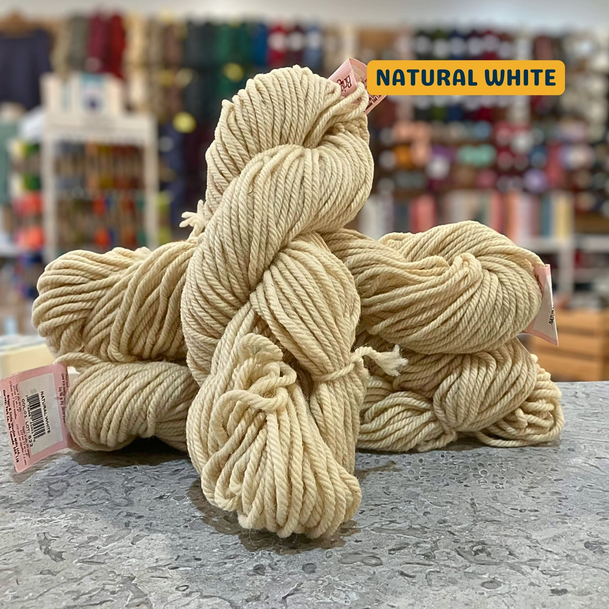 113g ecru (natural white) Canadian wool super bulky skein, Briggs & Little yarn, craft shop background (blurred)