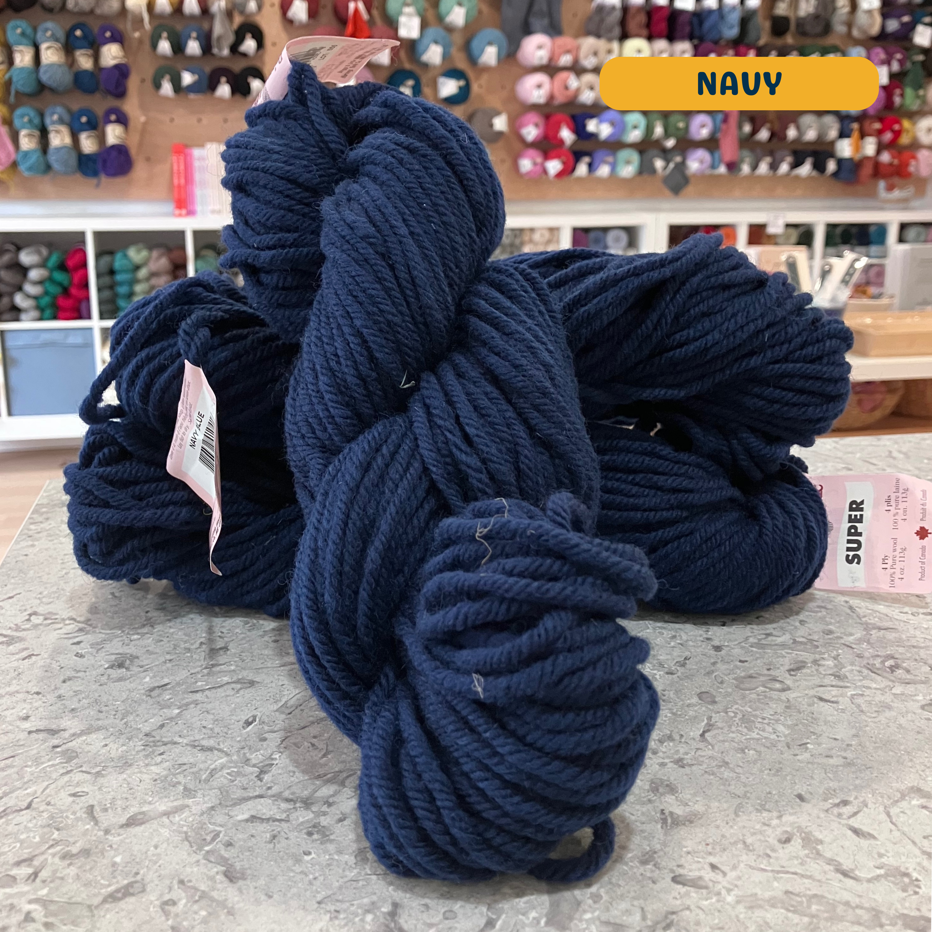 113g navy blue Canadian wool super bulky skein, Briggs & Little yarn, craft shop background (blurred)

