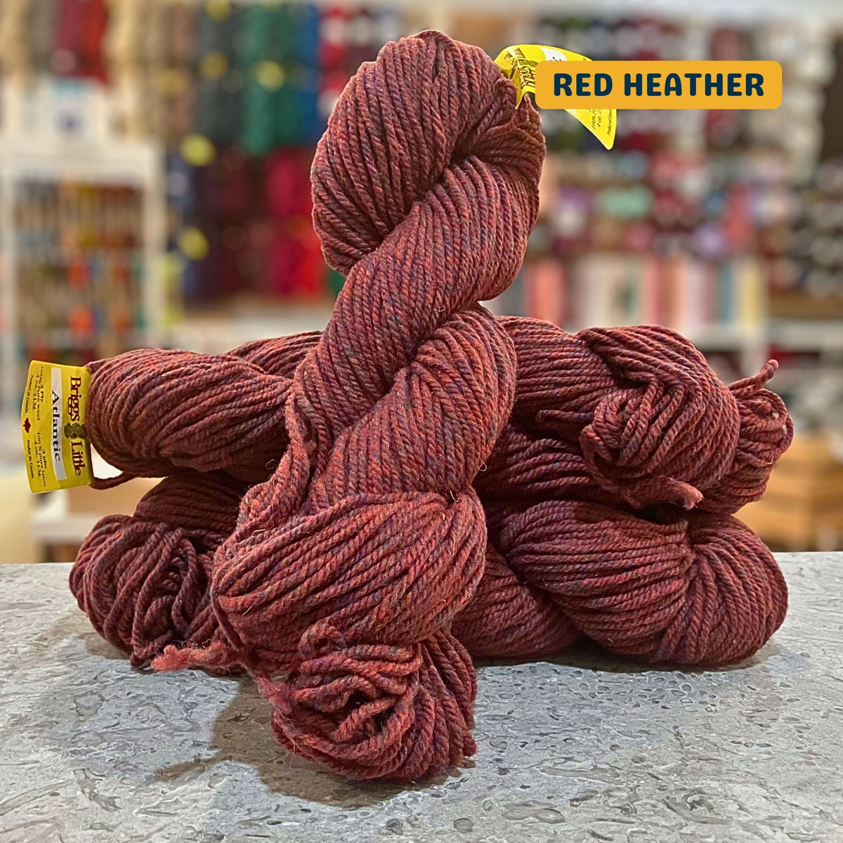 113g rust-red (red heather) Canadian wool bulky skein, Briggs & Little yarn, craft shop background (blurred)