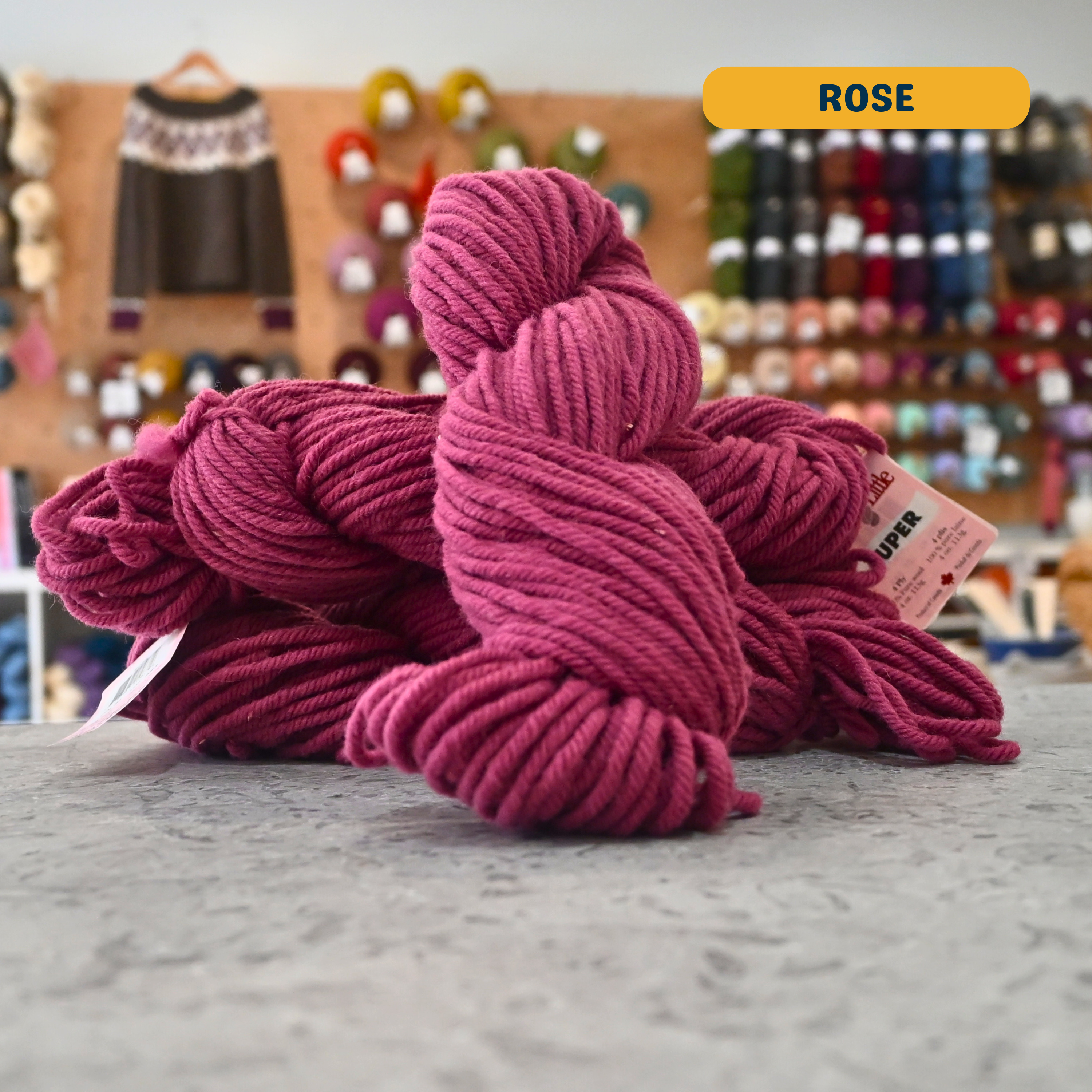 113g light maroon (rose) Canadian wool super bulky skein, Briggs & Little yarn, craft shop background (blurred)