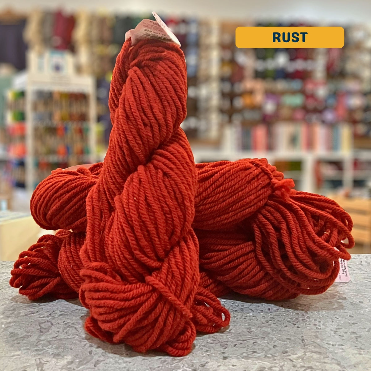 113g bright red-orange (rust) Canadian wool super bulky skein, Briggs & Little yarn, craft shop background (blurred)