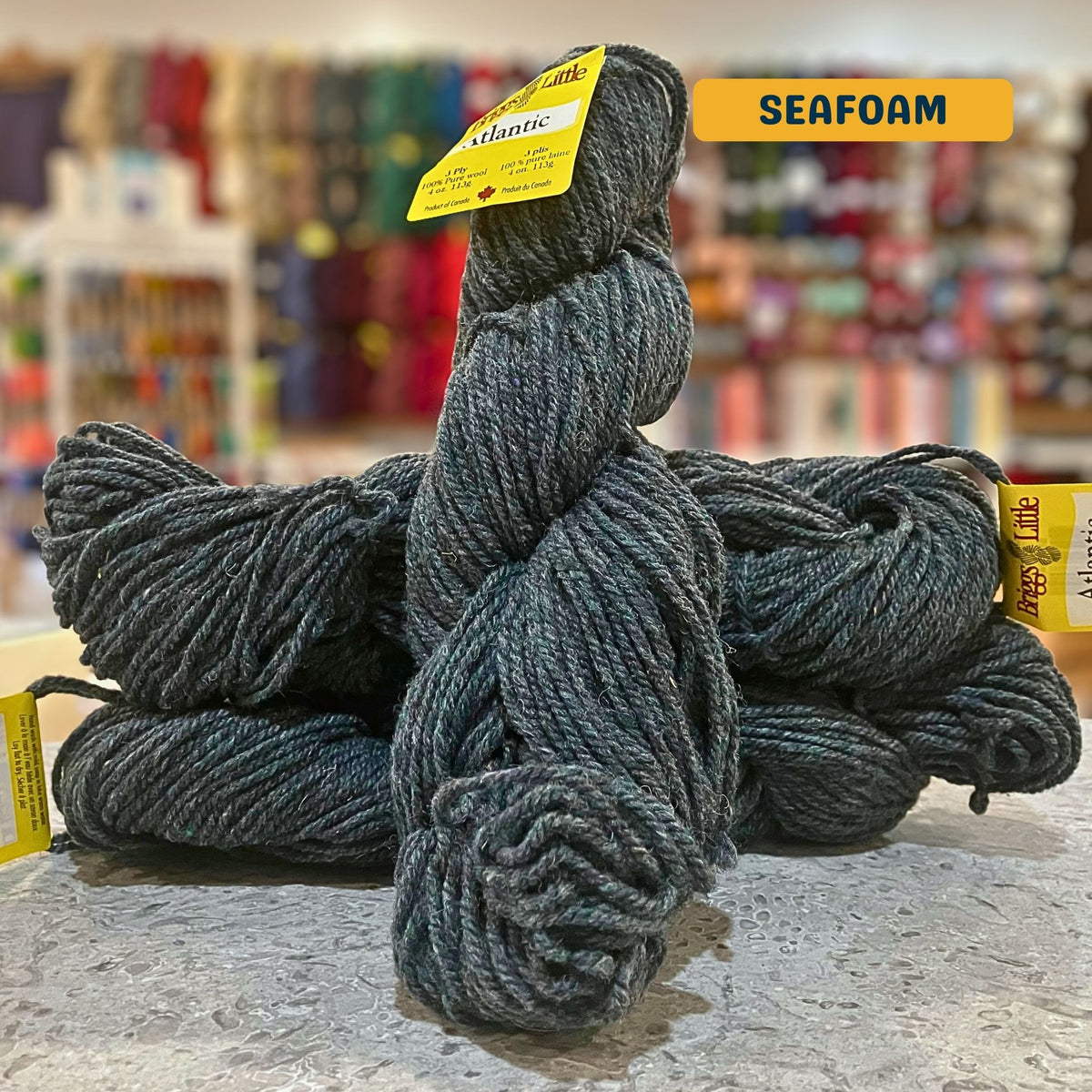 113g heathered grey-green (seafoam) Canadian wool bulky skein, Briggs & Little yarn, craft shop background (blurred)