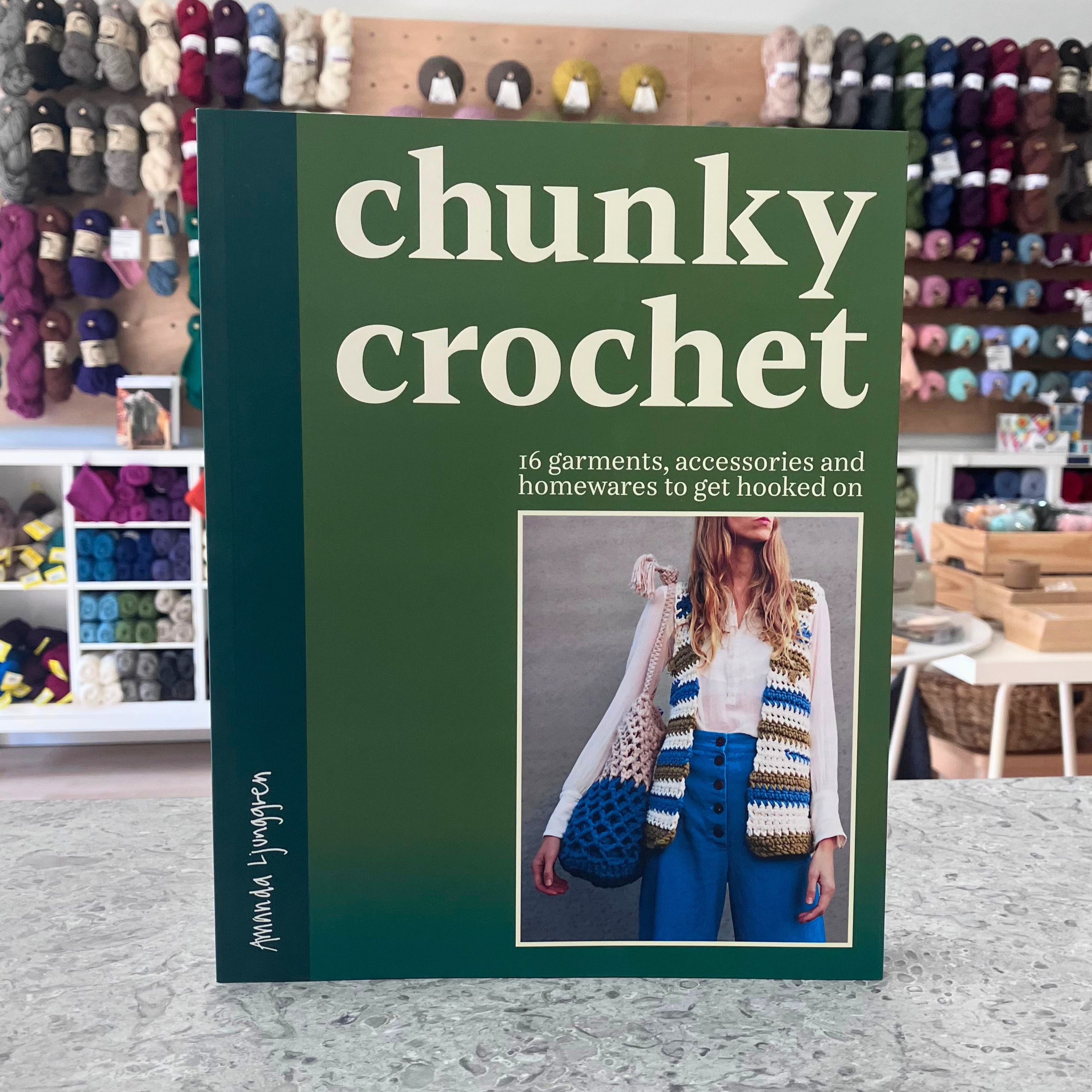 Chunky Crochet book by Amanda Ljunggren — cover showing chunky crocheted garments and homewares. Blurred craft shop background.