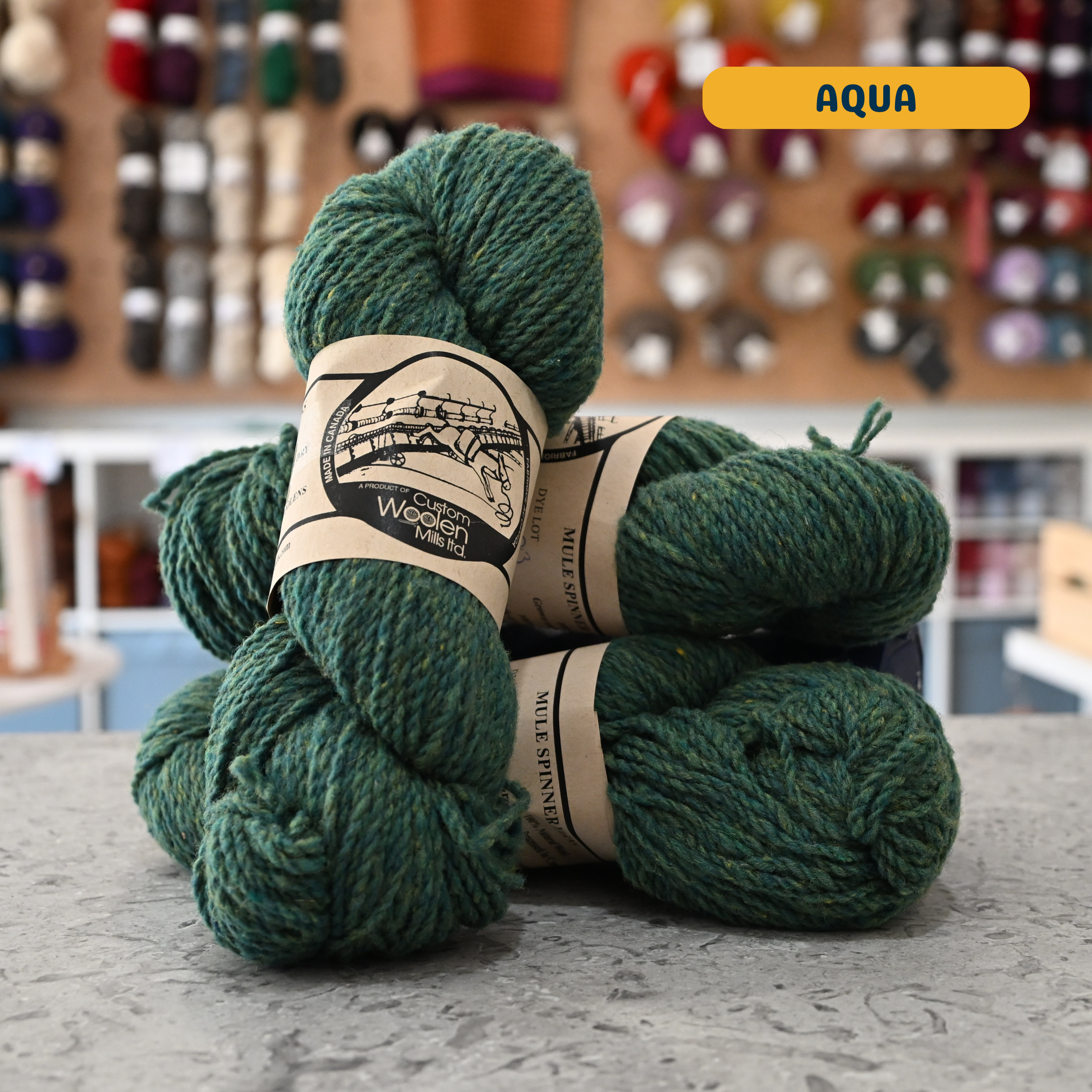 112g green (aqua) Canadian wool worsted yarn, 'Custom Woolen Mills' label visible, craft shop background (blurred)
