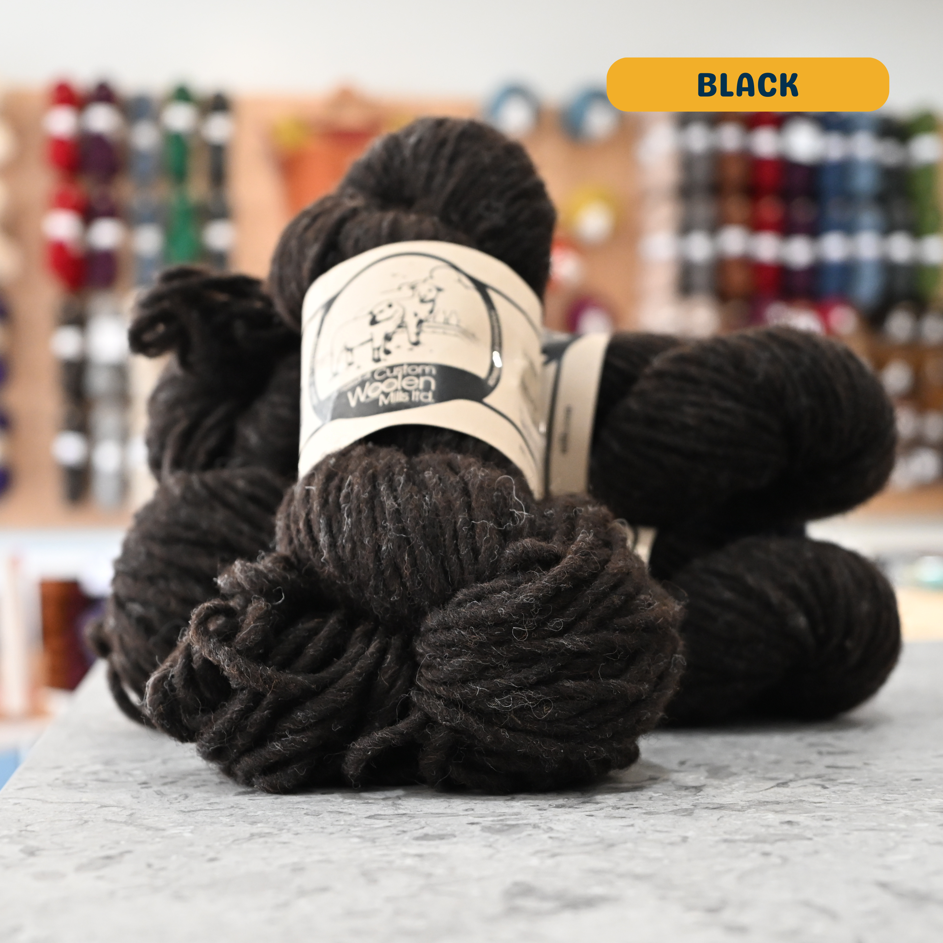 112g brown-black (black) Canadian wool bulky yarn, 'Custom Woolen Mills' label visible, craft shop background (blurred)
