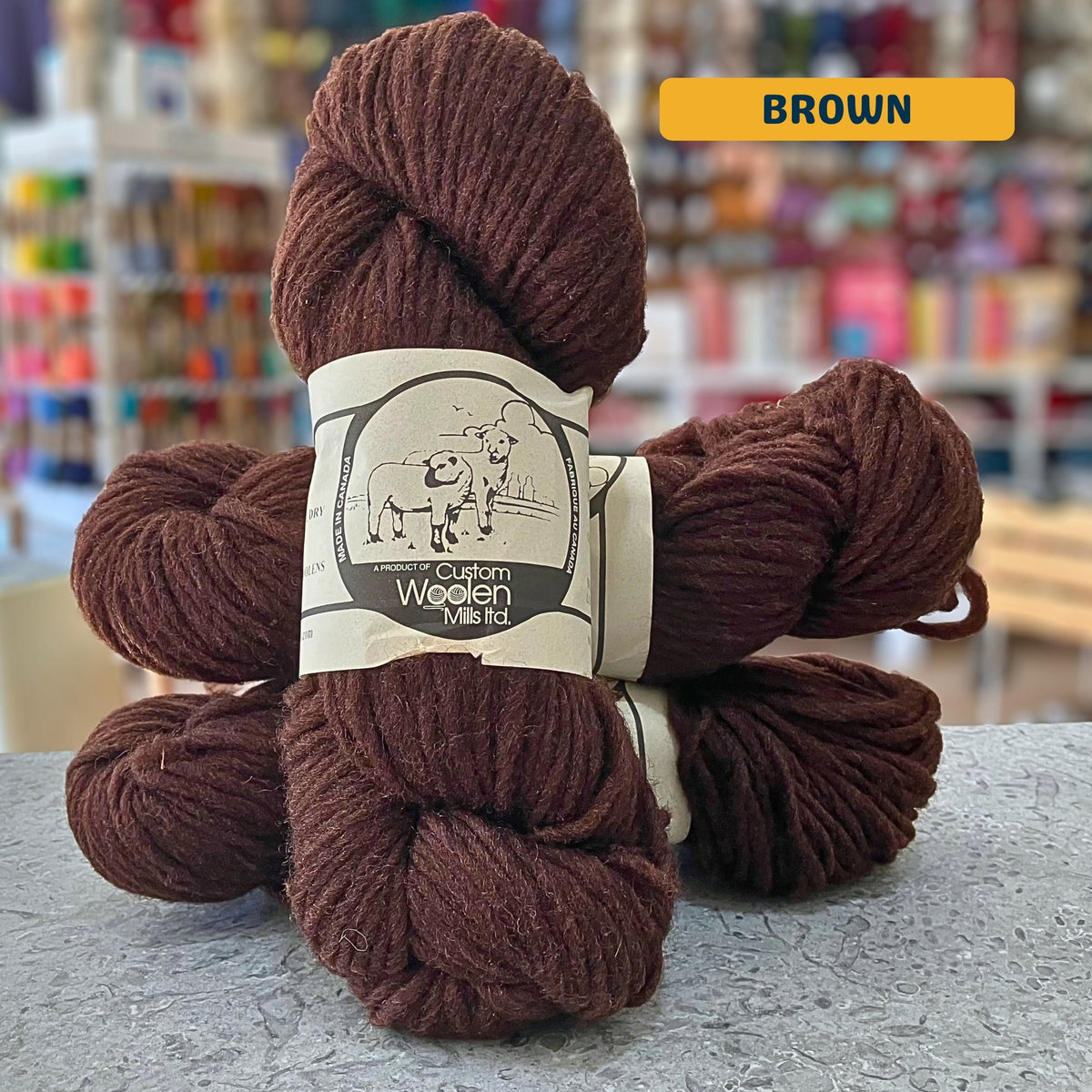 112g brown Canadian wool bulky yarn, 'Custom Woolen Mills' label visible, craft shop background (blurred)