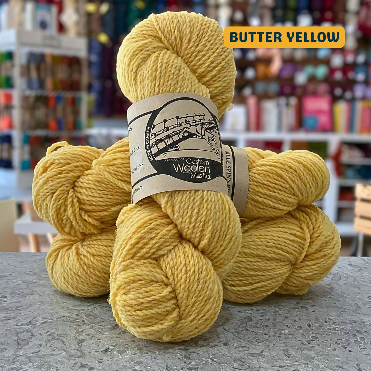 112g light yellow (butter yellow)  Canadian wool worsted yarn, 'Custom Woolen Mills' label visible, craft shop background (blurred)
