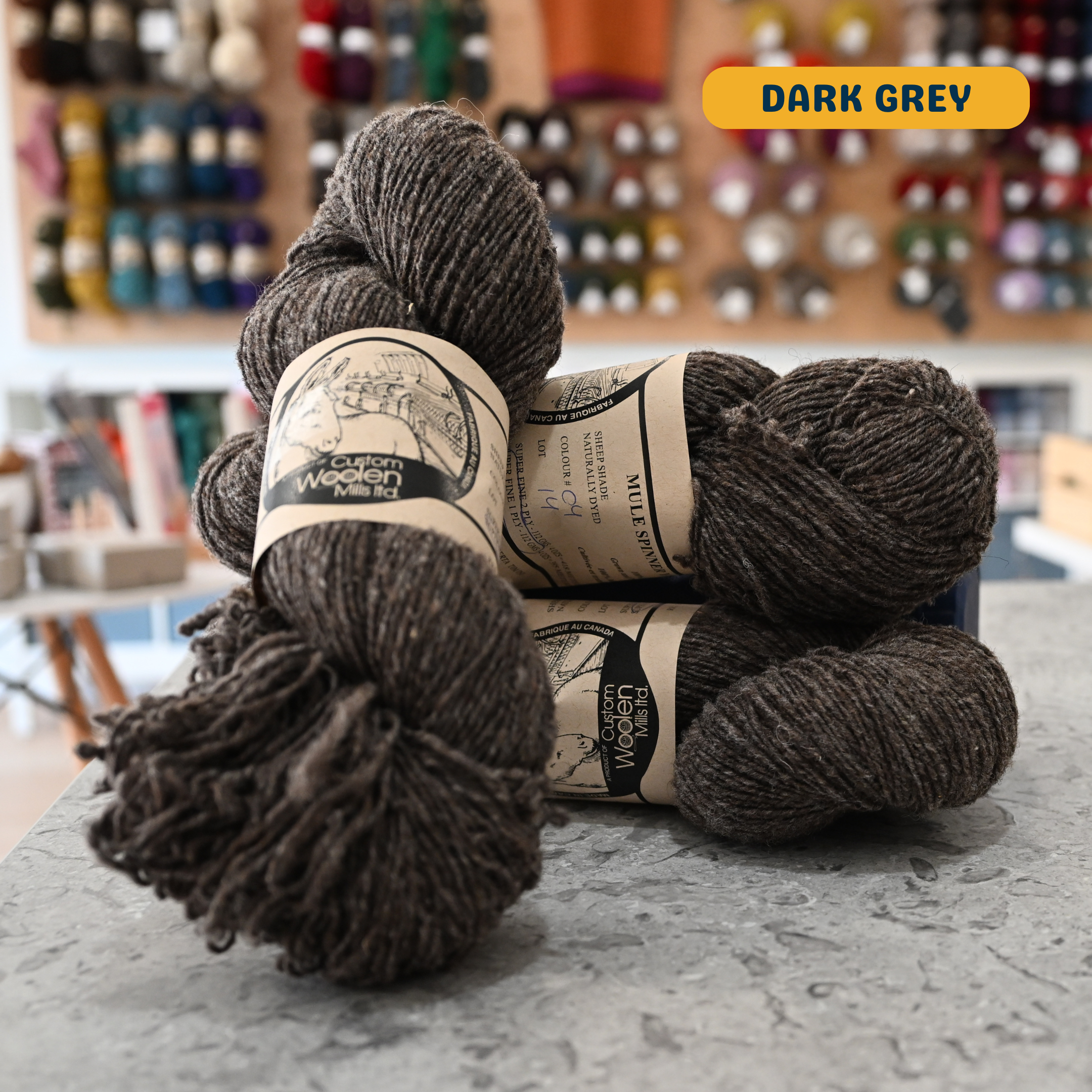 113g natural undyed dark grey Canadian wool fingering yarn, 'Custom Woolen Mills' label visible, craft shop background (blurred)