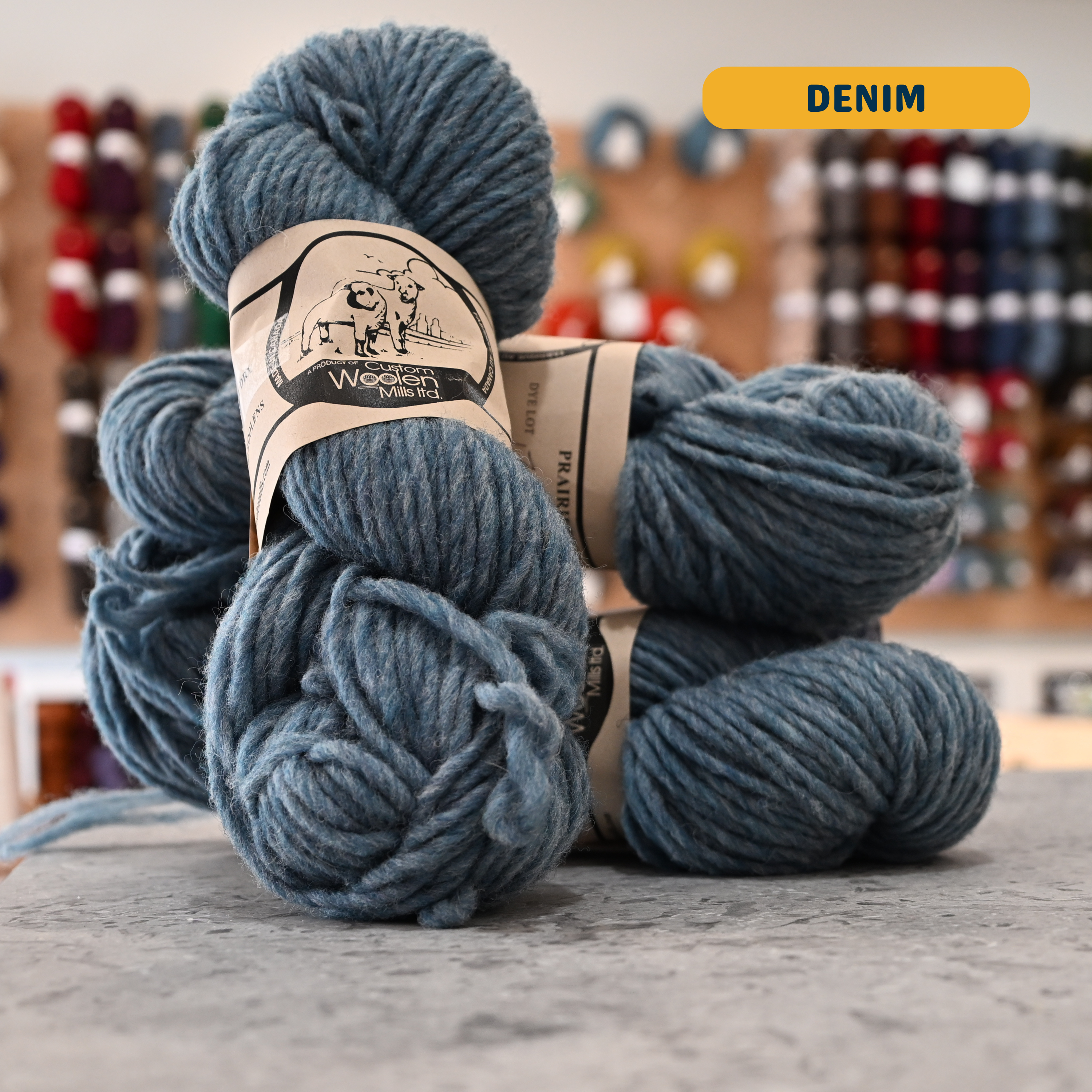 112g light blue (denim) Canadian wool bulky yarn, 'Custom Woolen Mills' label visible, craft shop background (blurred)
