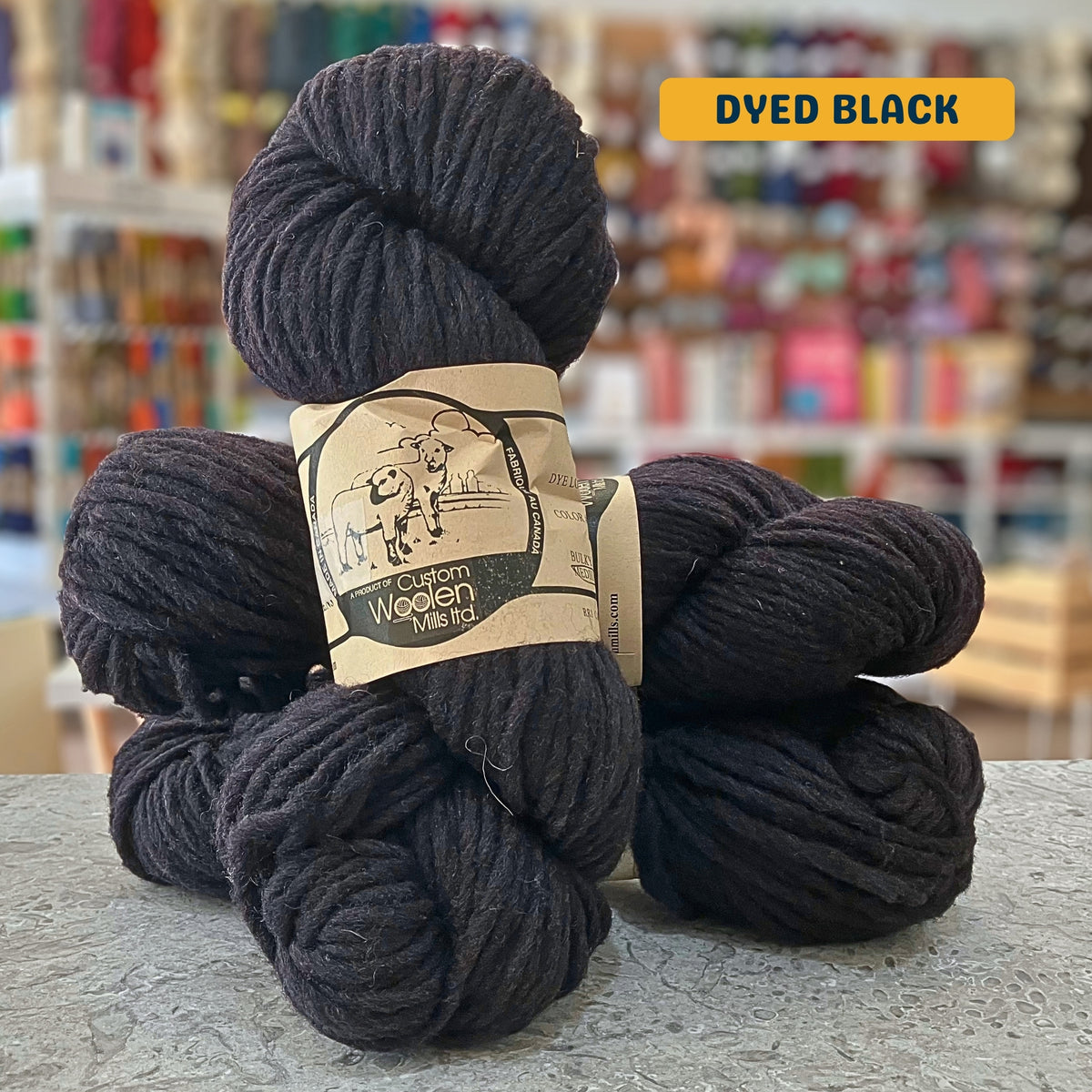 112g dyed black Canadian wool bulky yarn, 'Custom Woolen Mills' label visible, craft shop background (blurred)