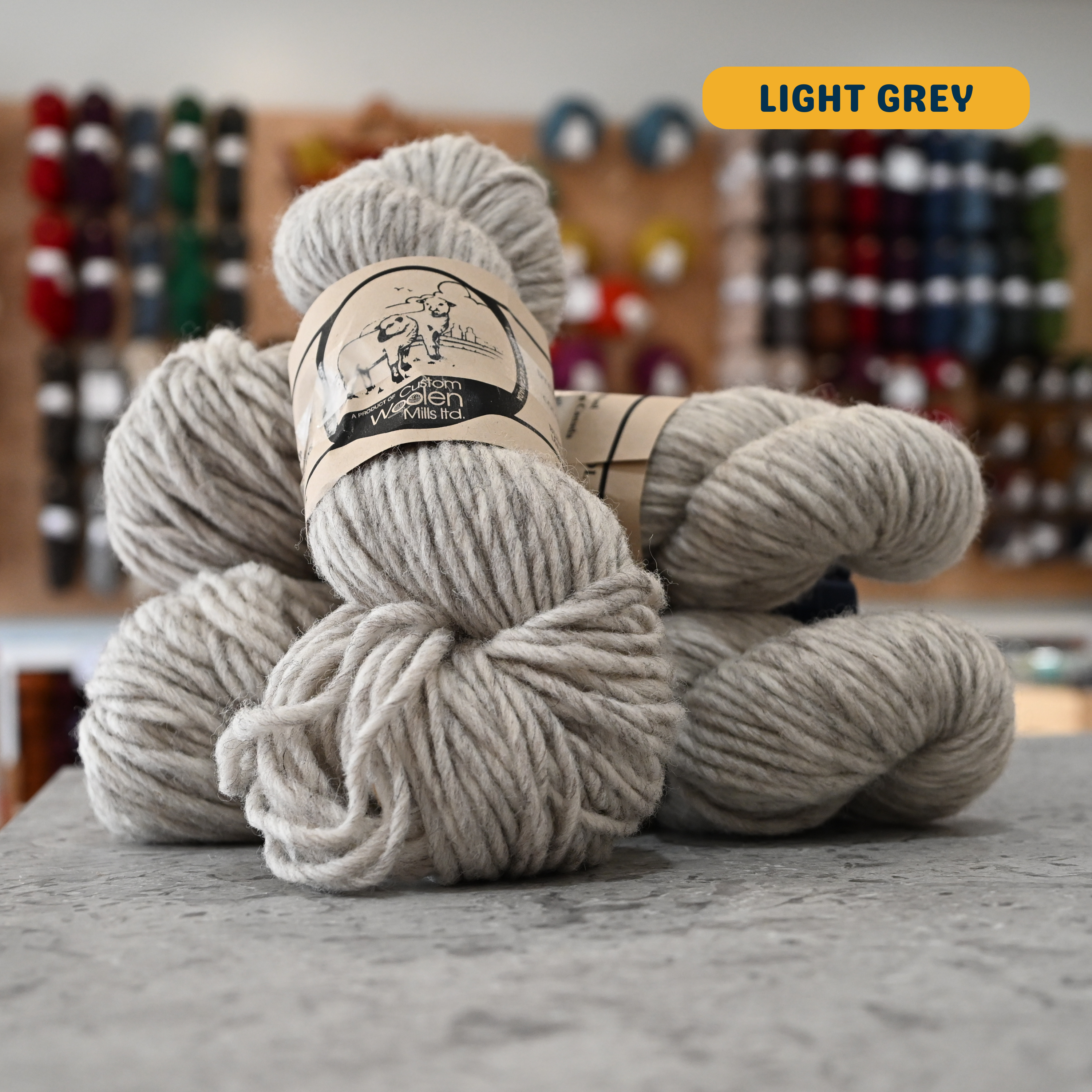 112g light grey Canadian wool bulky yarn, 'Custom Woolen Mills' label visible, craft shop background (blurred)
