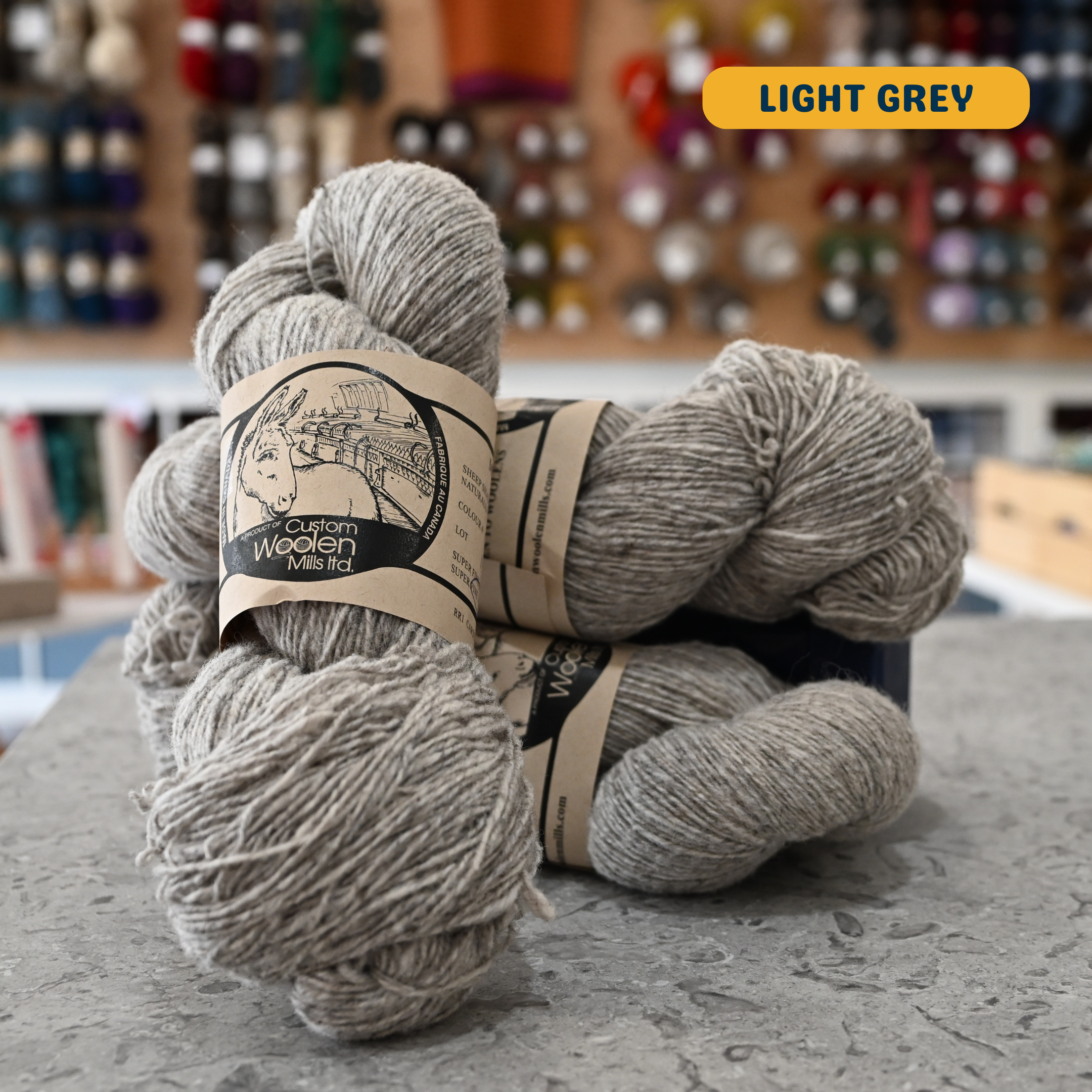 113g natural undyed light grey Canadian wool fingering yarn, 'Custom Woolen Mills' label visible, craft shop background (blurred)