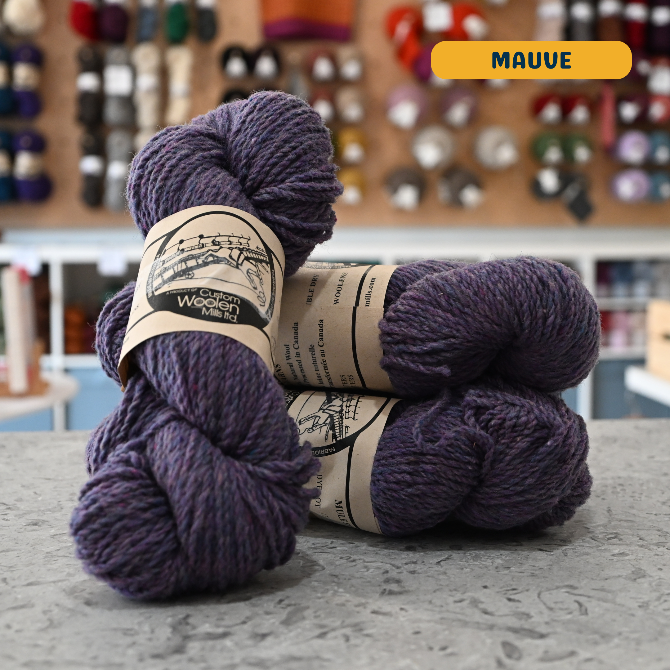 112g purple-grey (mauve) Canadian wool worsted yarn, 'Custom Woolen Mills' label visible, craft shop background (blurred)
