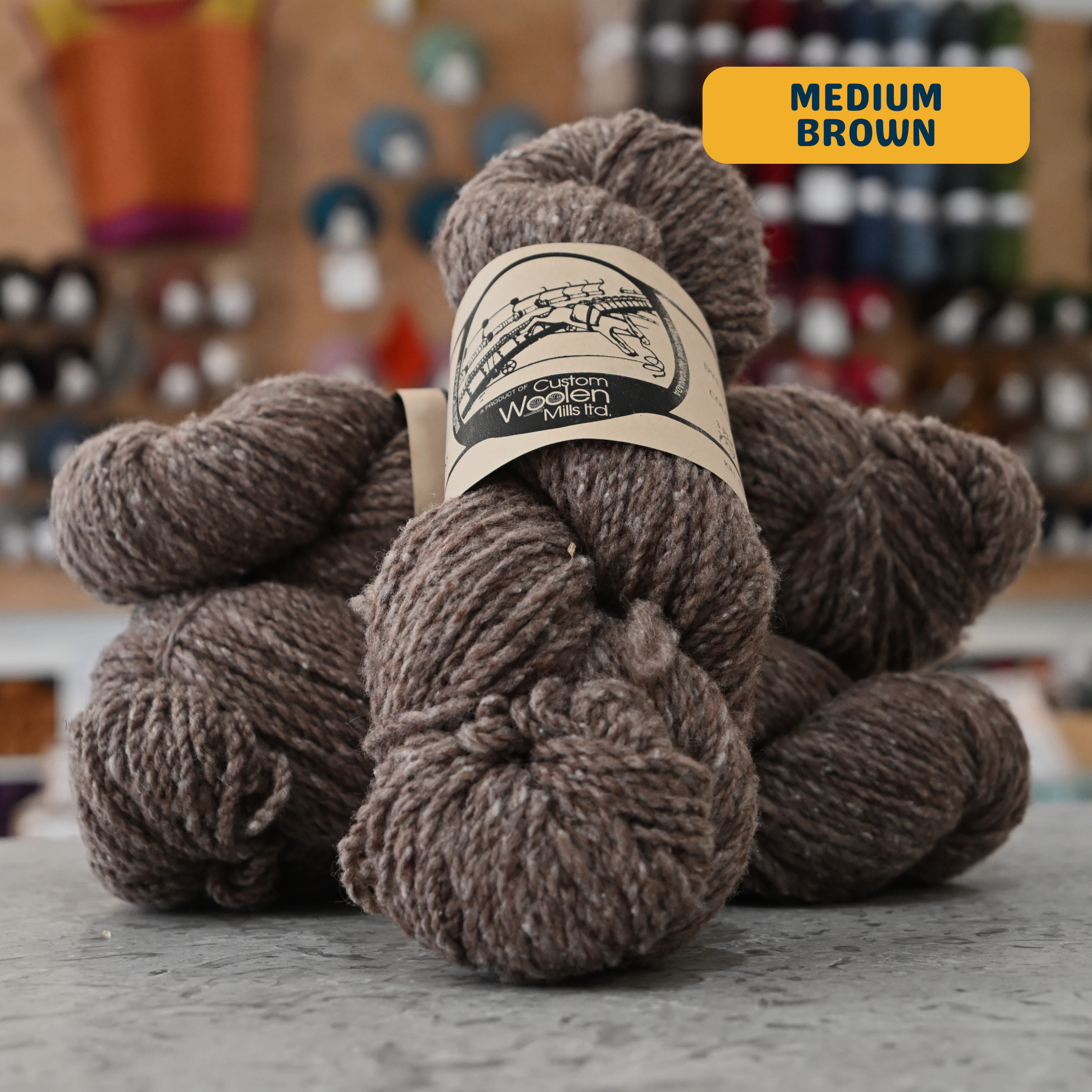 112g grey-brown (medium brown) Canadian wool worsted yarn, 'Custom Woolen Mills' label visible, craft shop background (blurred)