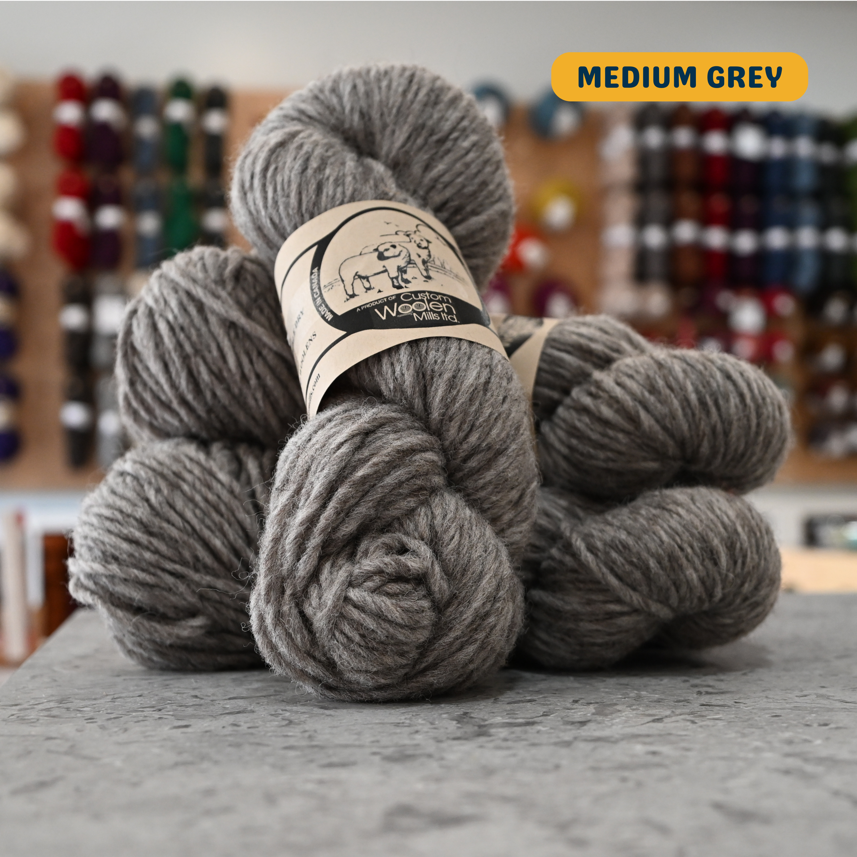 112g medium grey Canadian wool bulky yarn, 'Custom Woolen Mills' label visible, craft shop background (blurred)