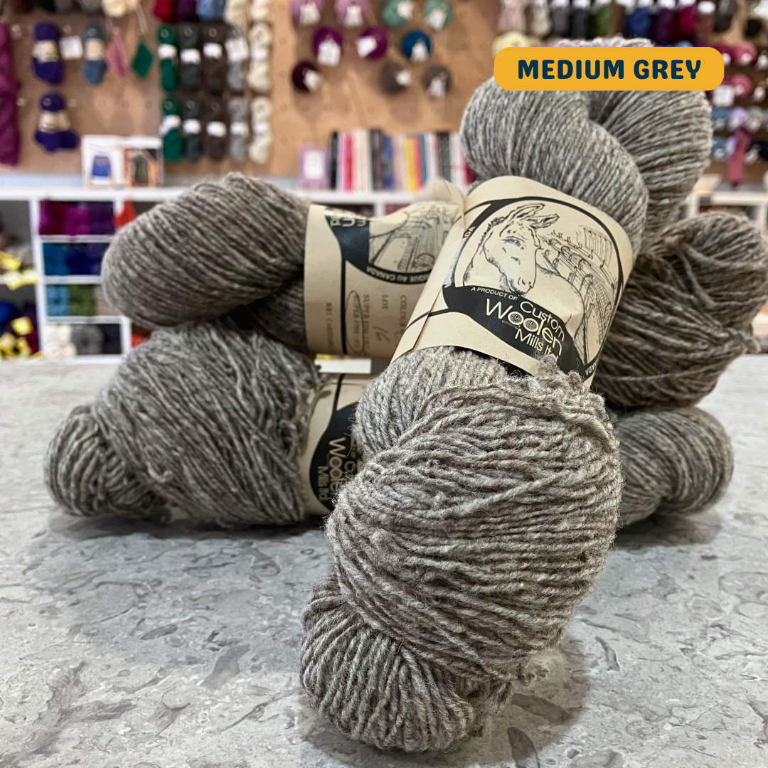 113g natural undyed medium grey Canadian wool fingering yarn, 'Custom Woolen Mills' label visible, craft shop background (blurred)
