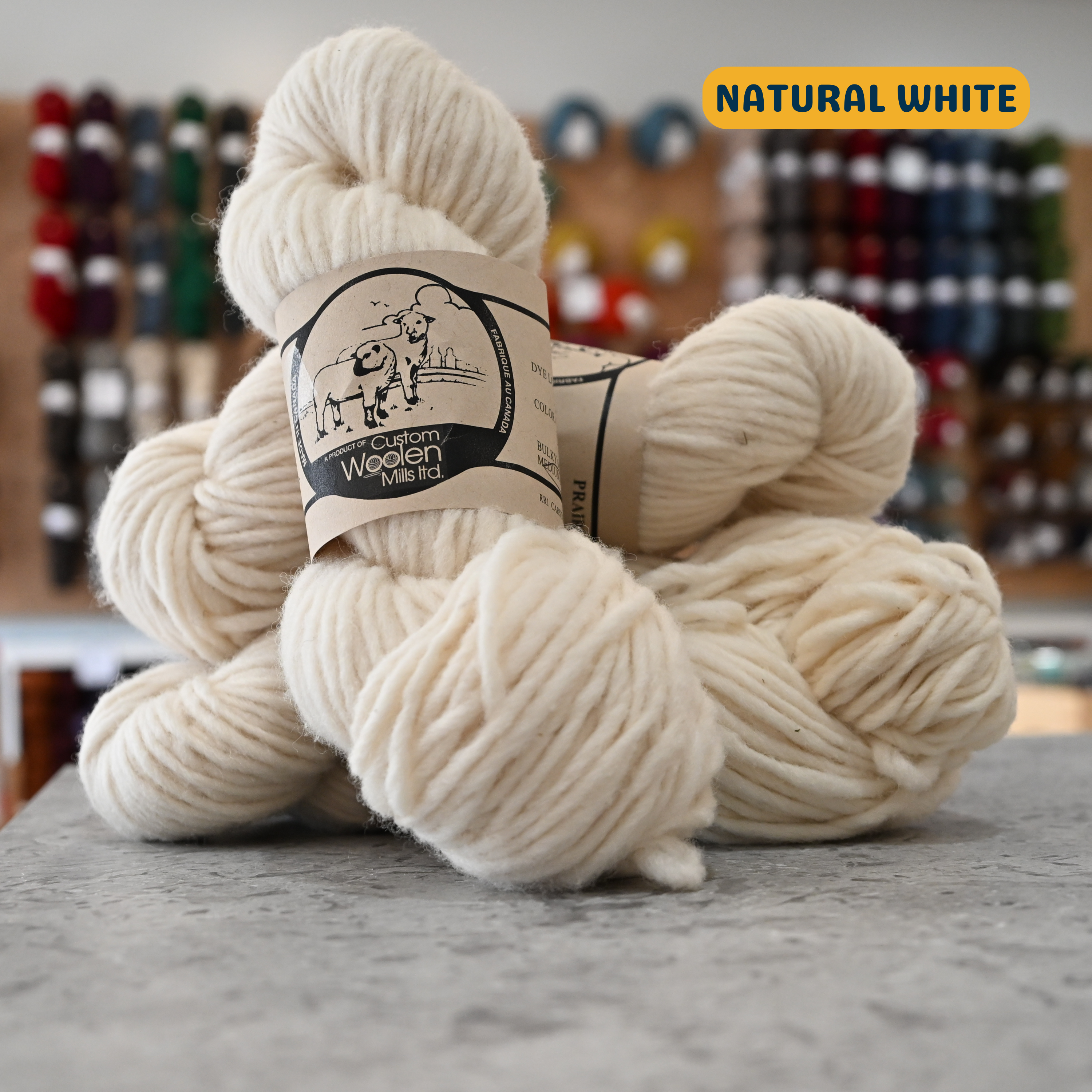 112g natural white Canadian wool bulky yarn, 'Custom Woolen Mills' label visible, craft shop background (blurred)
