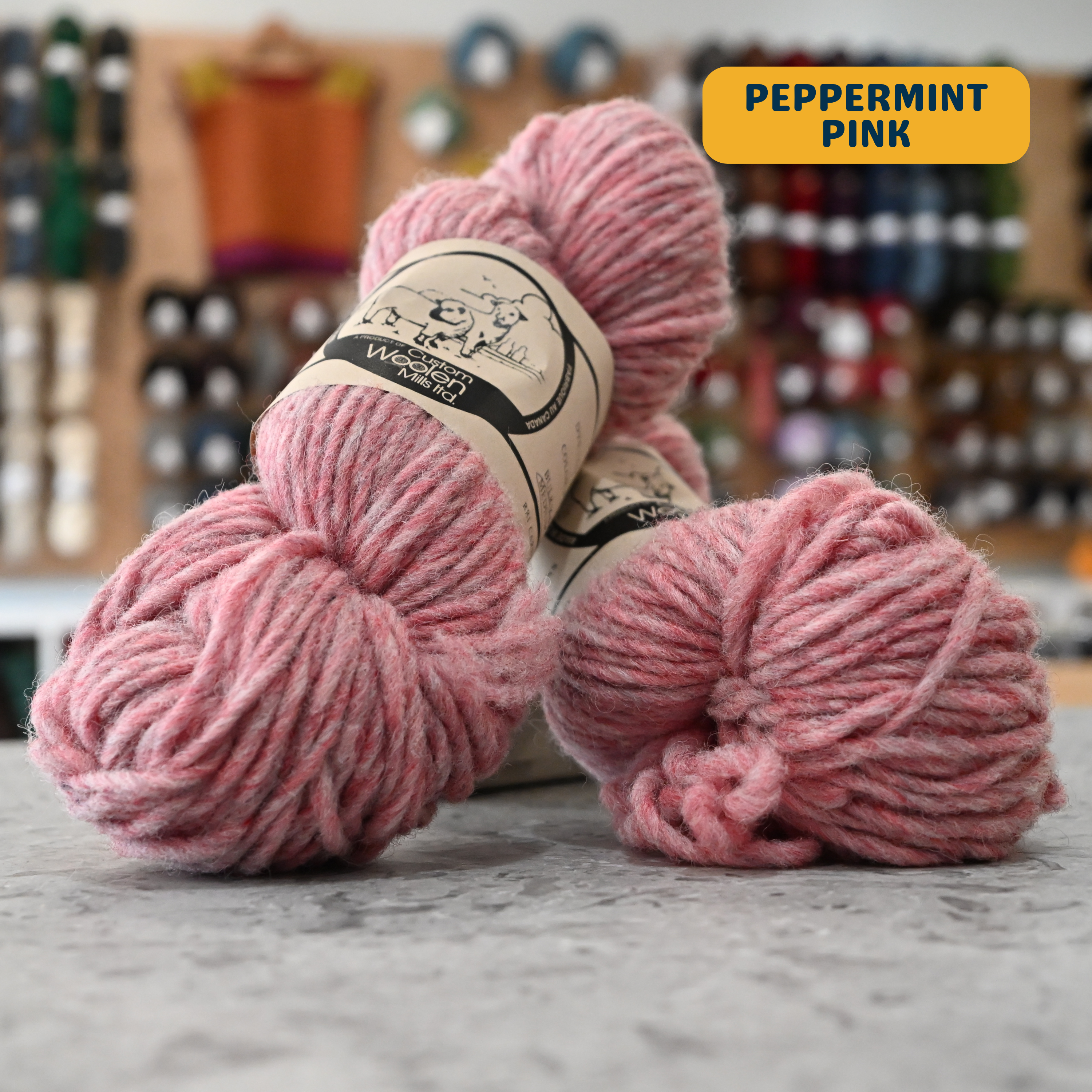 112g light pink (peppermint pink) Canadian wool bulky yarn, 'Custom Woolen Mills' label visible, craft shop background (blurred)
