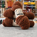 112g orange-brown (rust) Canadian wool bulky yarn, 'Custom Woolen Mills' label visible, craft shop background (blurred)
