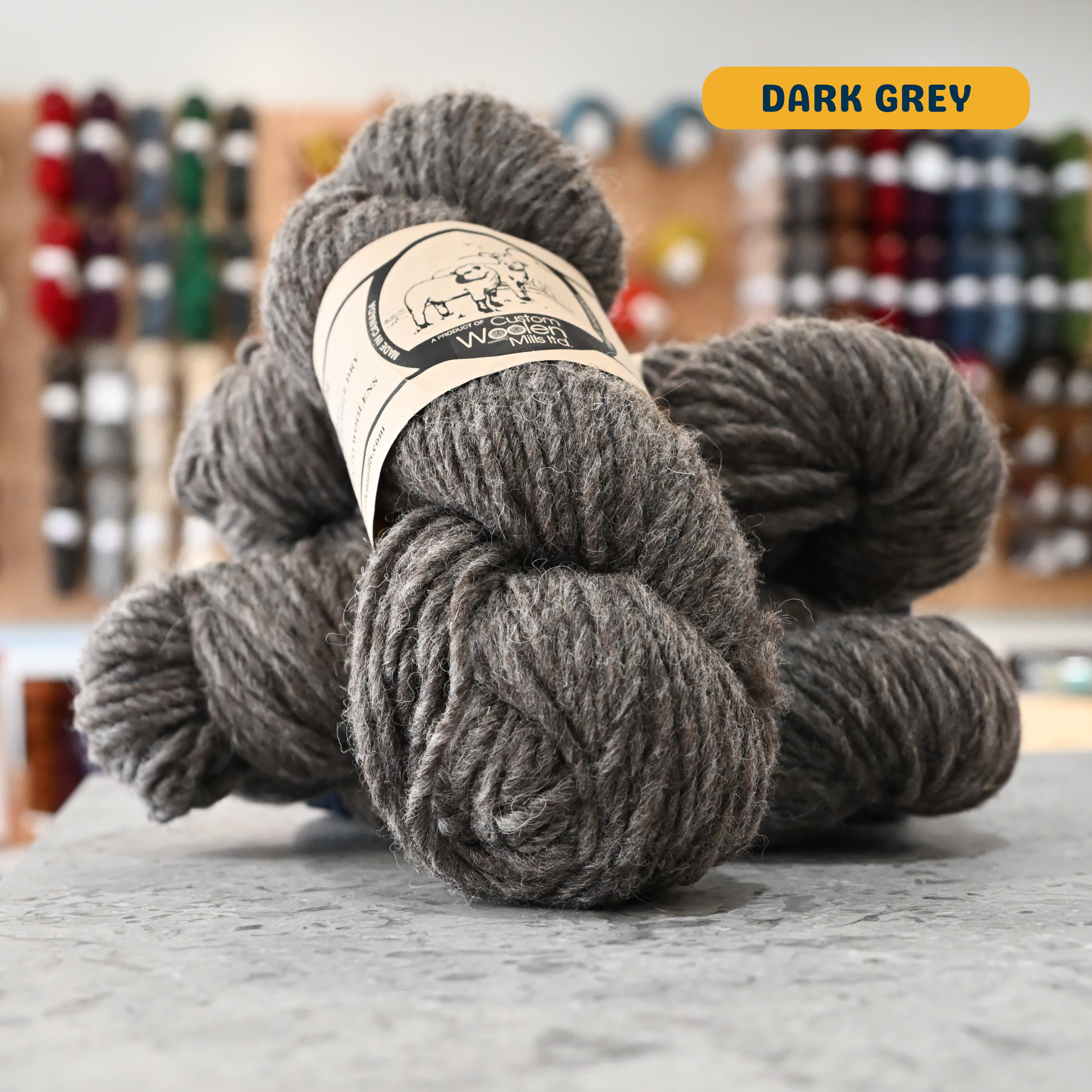 112g dark grey Canadian wool bulky yarn, 'Custom Woolen Mills' label visible, craft shop background (blurred)
