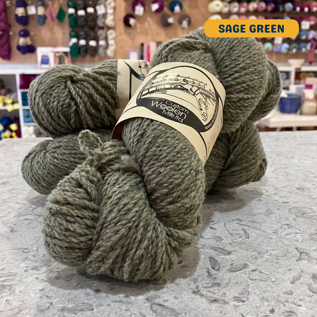 112g sage green Canadian wool worsted yarn, 'Custom Woolen Mills' label visible, craft shop background (blurred)
