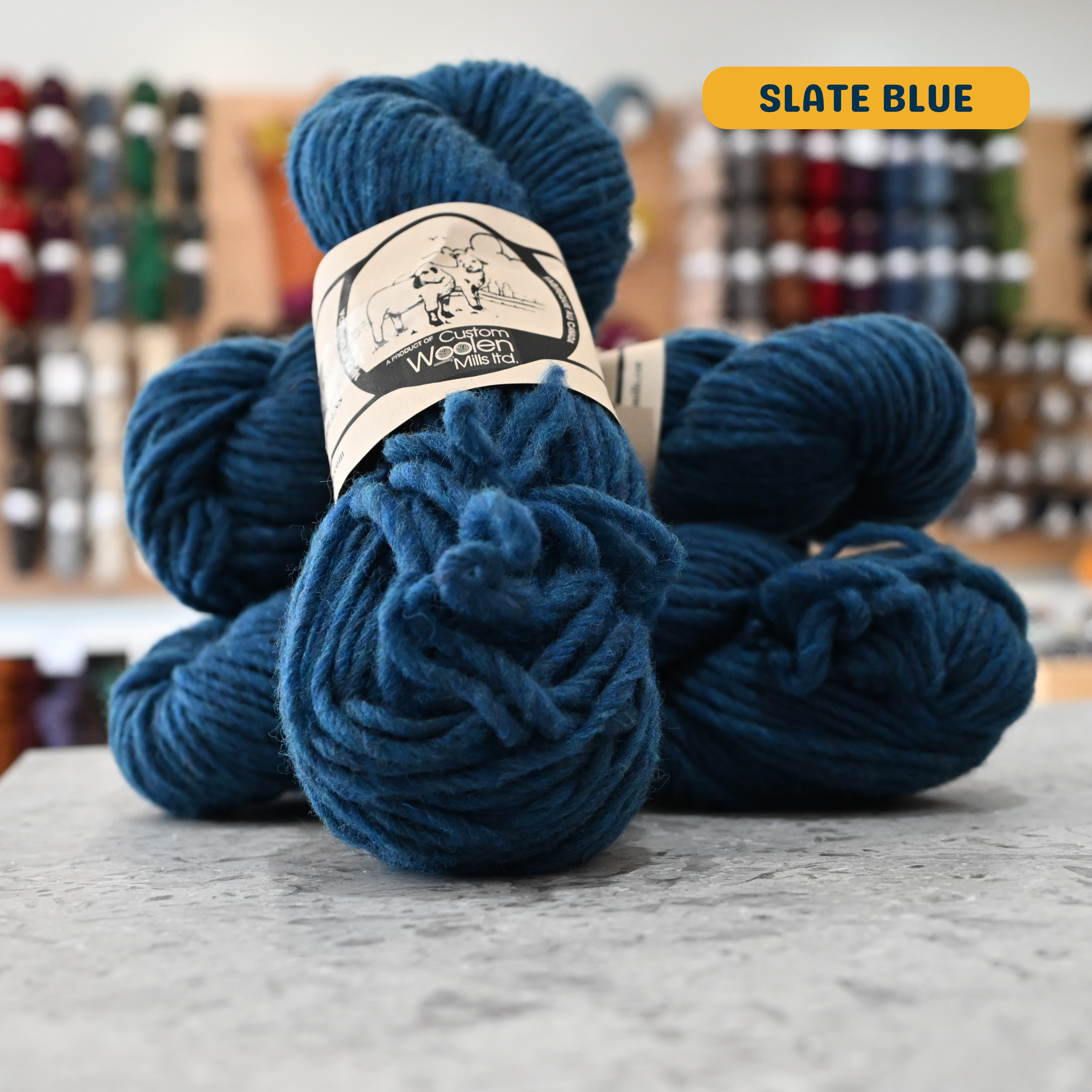112g slate blue Canadian wool bulky yarn, 'Custom Woolen Mills' label visible, craft shop background (blurred)