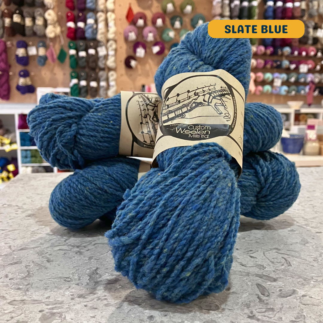 112g slate blue Canadian wool worsted yarn, 'Custom Woolen Mills' label visible, craft shop background (blurred)
