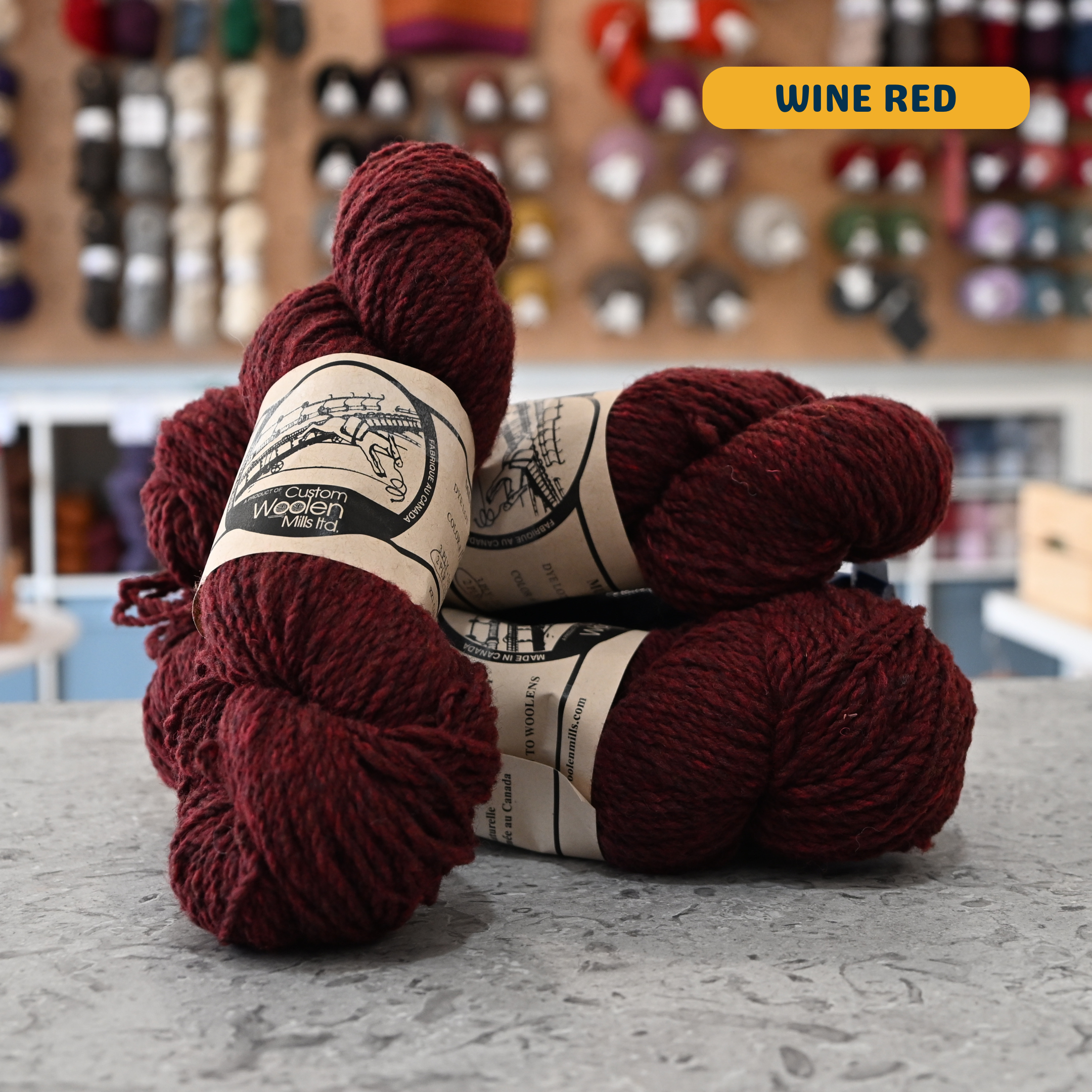112g wine red Canadian wool worsted yarn, 'Custom Woolen Mills' label visible, craft shop background (blurred)